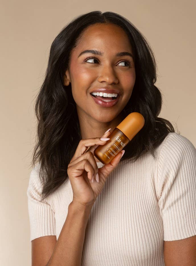 A smiling woman with long dark hair holds a small amber of Kitsch hair perfume against a warm beige background. 