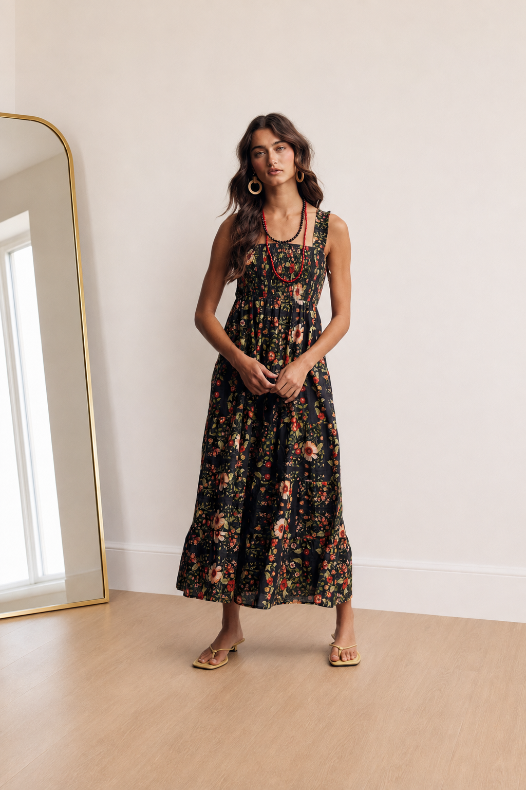 A woman stands in a bright room wearing a sleeveless midi dress with a dense floral print on a black background. The dress features a scoop neckline, thin shoulder straps, and a gathered waist.