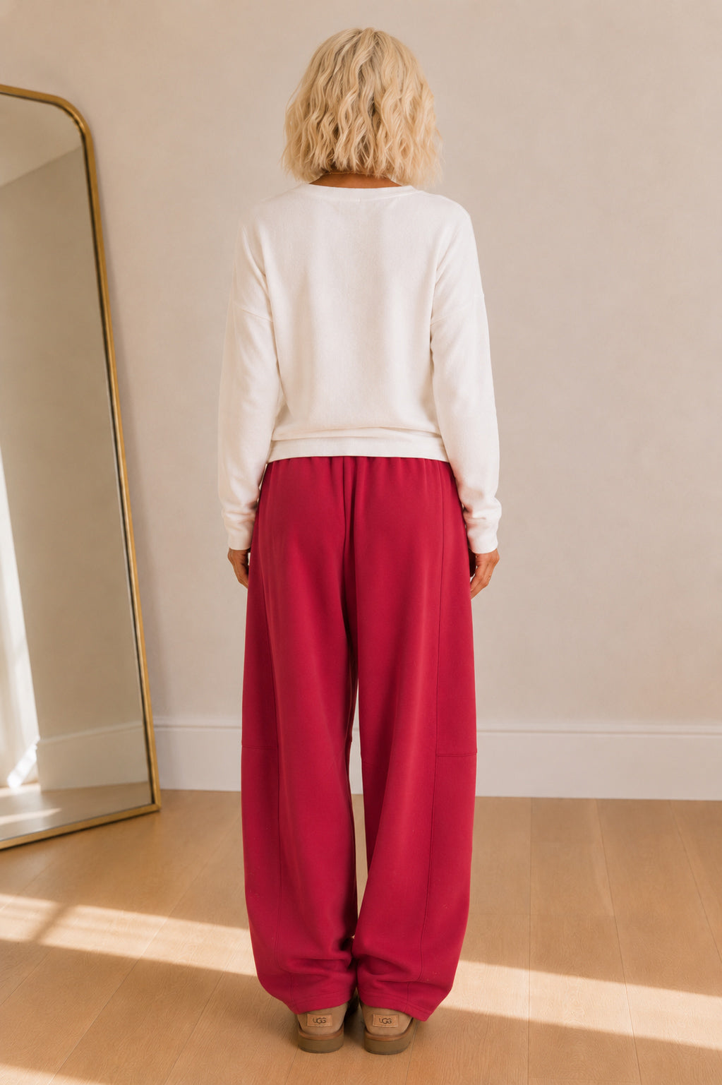 A woman viewed from behind wearing a white long-sleeve top and bright pink wide-leg pants, standing in a sunlit room with a wooden floor and a mirror on the left.
