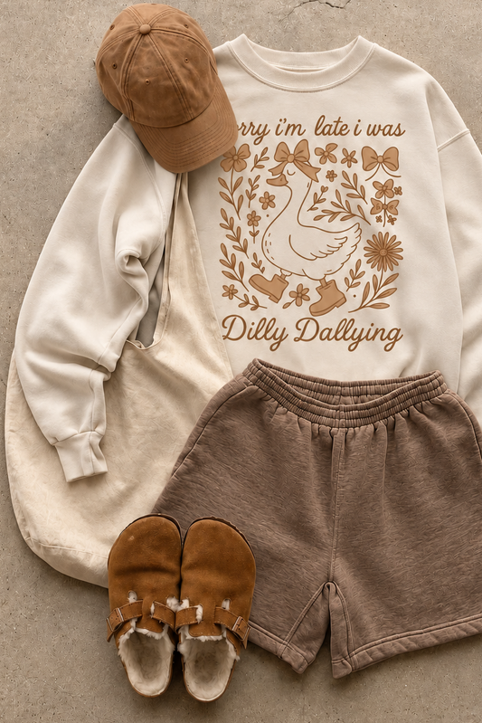 A styled flat-lay of a cream sweatshirt with a brown duck graphic and the text 'Dilly Dallying,' paired with brown shorts, a tan cap, and tan slip-on shoes on a concrete surface.
