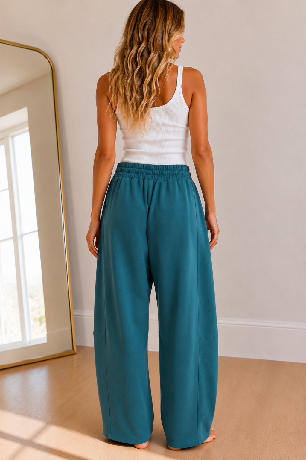 A woman shown from behind wearing wide-leg teal pants and a white tank top in a bright room with a mirror and light wood floor.