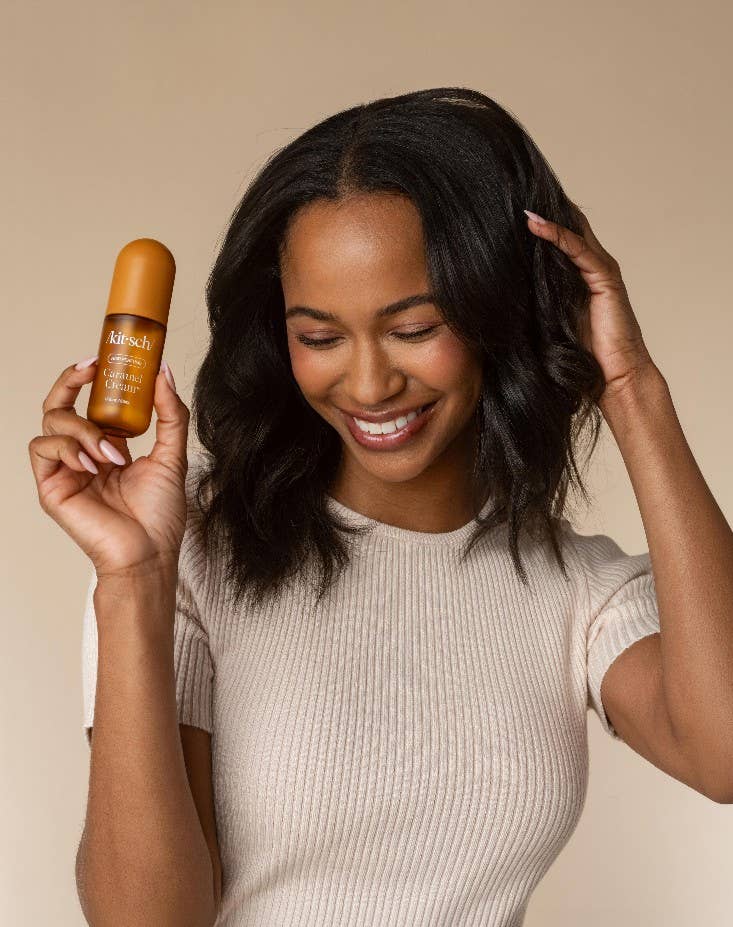 A woman with dark wavy hair smiles while holding a small amber bottle labeled kitsch hair perfume in her right hand, against a plain beige studio background.