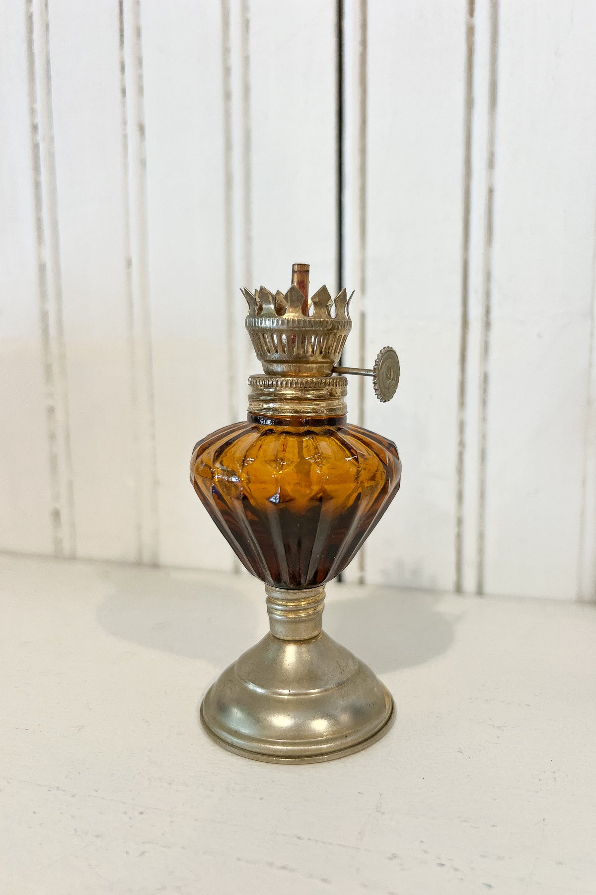 A vintage-style oil lamp with a faceted amber glass body and a metallic brass-toned base, collar, and wick holder, photographed against a white paneled wall.
