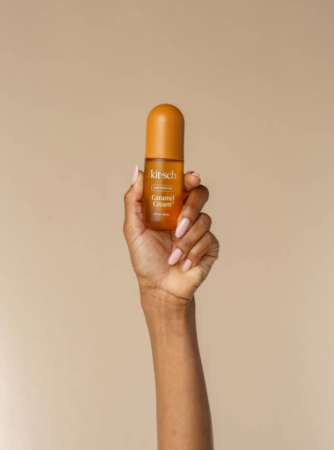 A hand with light pink manicured nails holds a small amber bottle of kitsch Caramel cream hair perfume in front of a solid beige background.