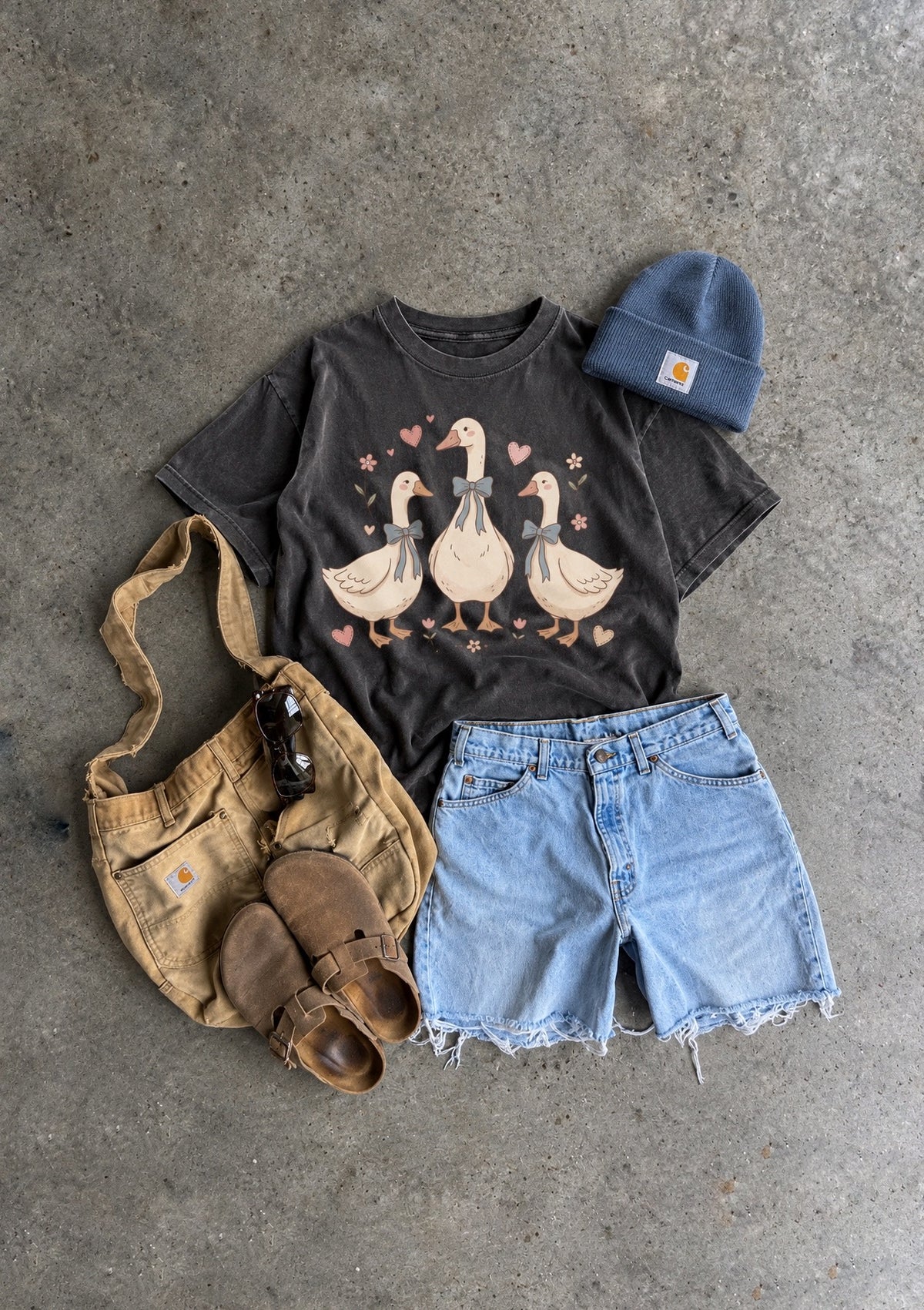 A styled flat-lay outfit on a gray concrete background featuring a dark charcoal graphic t-shirt with three ducks, light-wash denim shorts, a tan canvas shoulder bag, brown leather sandals, and a blue beanie. The t-shirt has a visible Carhartt logo on the beanie.