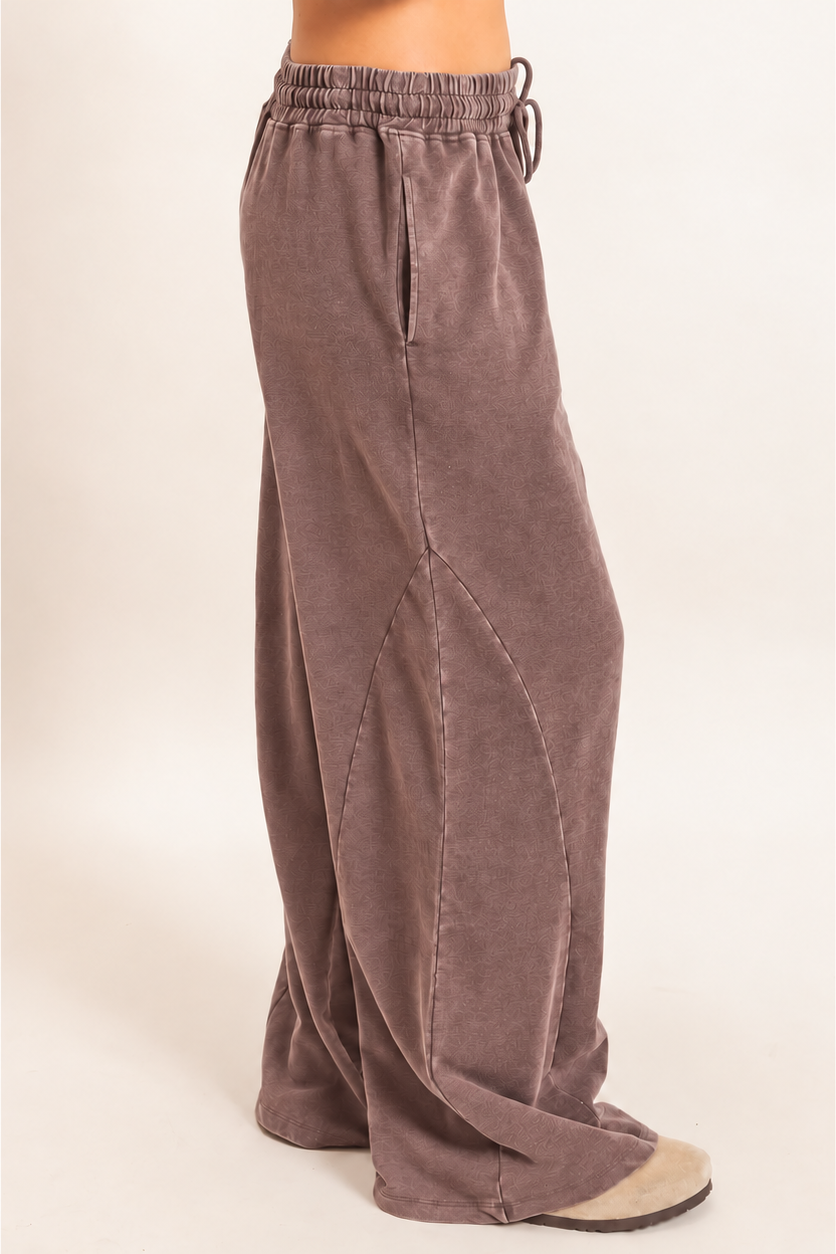A person wearing loose, wide-leg lounge pants in a muted taupe-brown color, shown from the waist down against a plain light beige studio background.