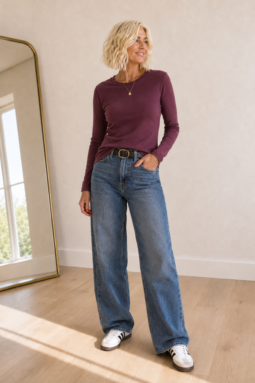 A woman modeling a long-sleeve crewneck top in deep plum purple, paired with wide-leg blue jeans and white sneakers, standing in a brightly lit room with a wooden floor and a large mirror.