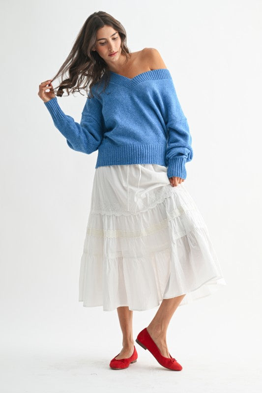 Full-length image of woman wearing a cozy blue sweater off one shoulder with a white tiered skirt and red flats, in a relaxed cottagecore pose.
