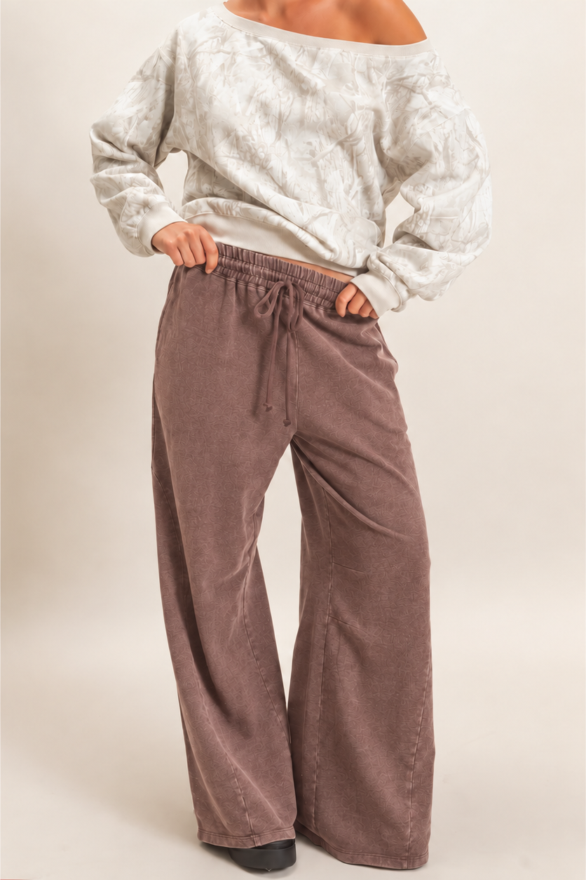 A model wearing a pair of wide-leg, dusty brown corduroy pants with a drawstring waist, paired with a light off-the-shoulder top, against a plain beige studio background.