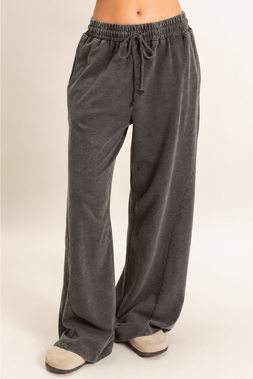 A person wearing dark gray wide-leg lounge pants with an elastic drawstring waist, paired with beige slip-on shoes, shown against a plain light beige studio background.