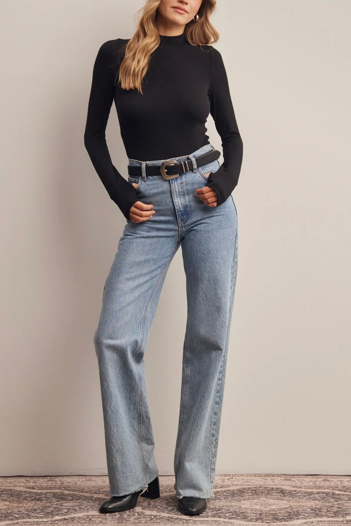 A woman modeling a black long-sleeve mock neck top and light-wash blue straight-leg jeans with a black belt, standing against a neutral beige wall.