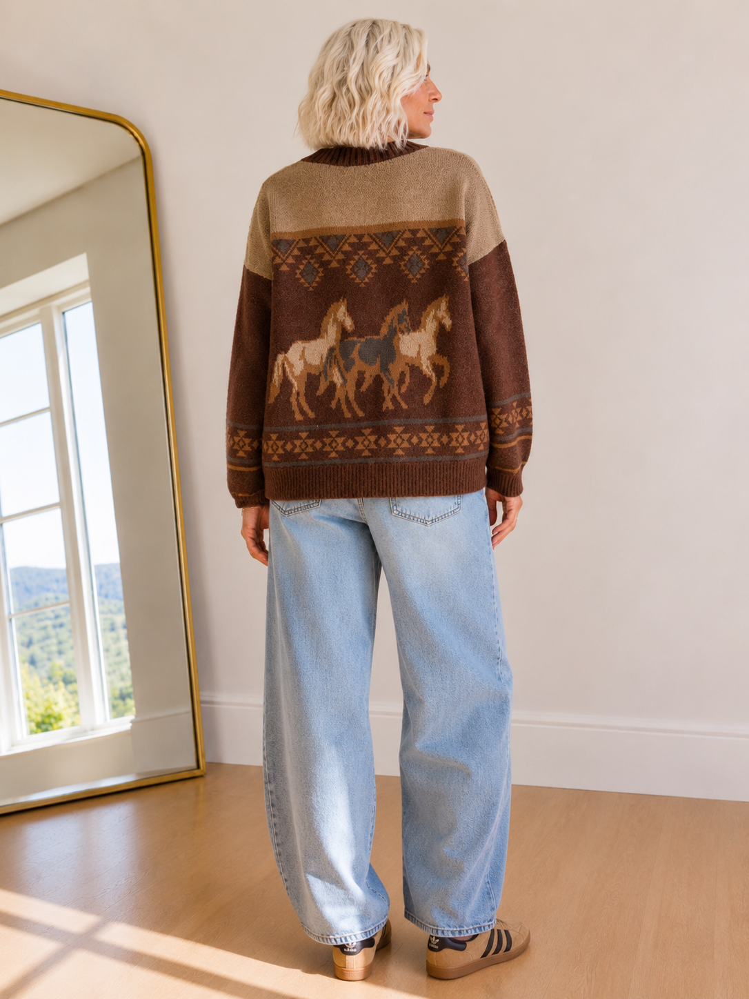 A woman stands with her back to the camera wearing a brown and beige knit sweater with a horse pattern, light blue jeans, and brown sneakers in a bright room with a large mirror.