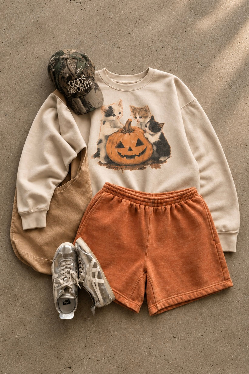 Fall outfit flat lay with a cream kitten Halloween sweatshirt, pumpkin-orange shorts, camouflage hat, tan tote and sneakers.