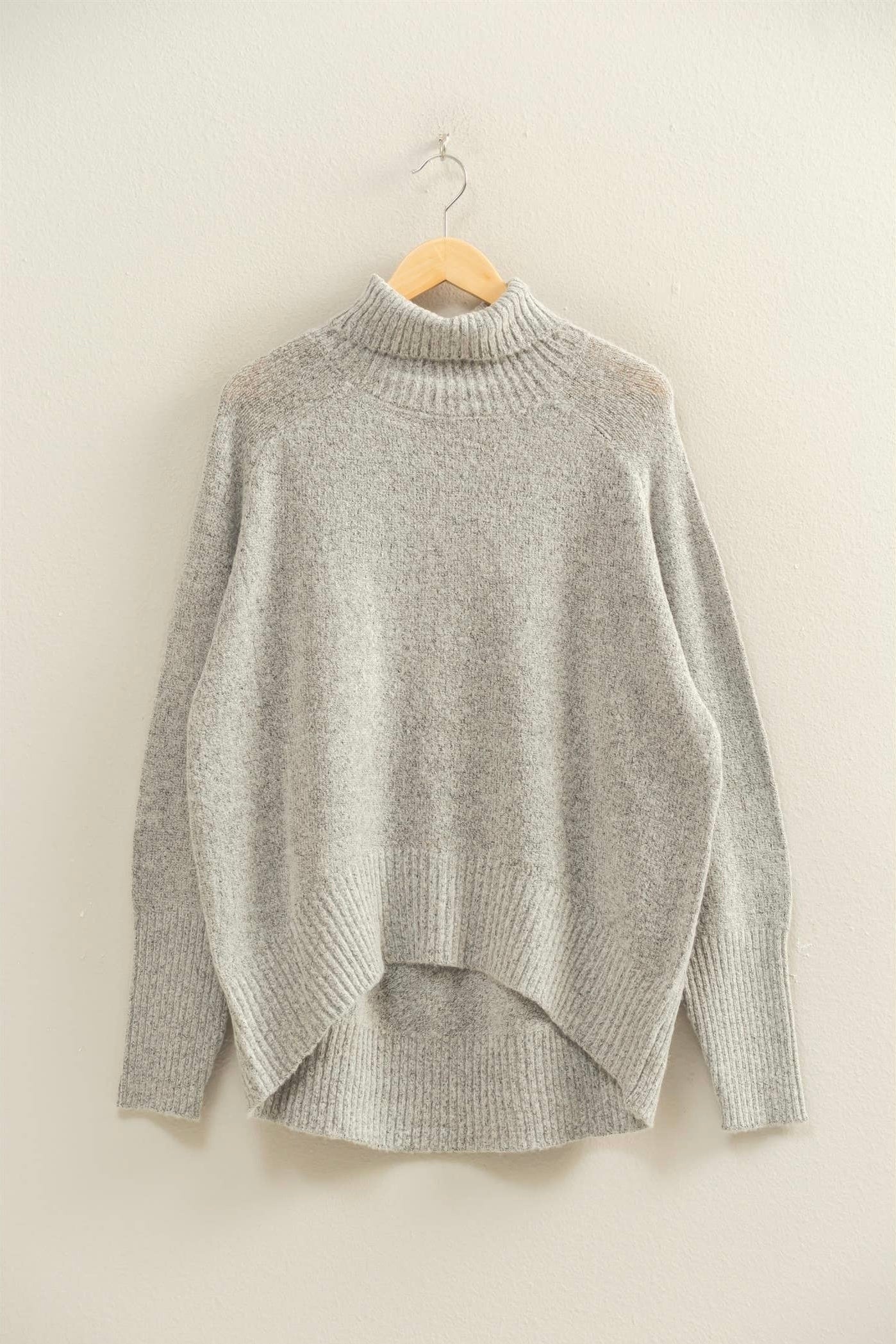 Heather grey mock-neck sweater hanging on a wooden hanger against a neutral wall. Ribbed neckline, cuffs, and hem with a relaxed silhouette and soft knit texture.