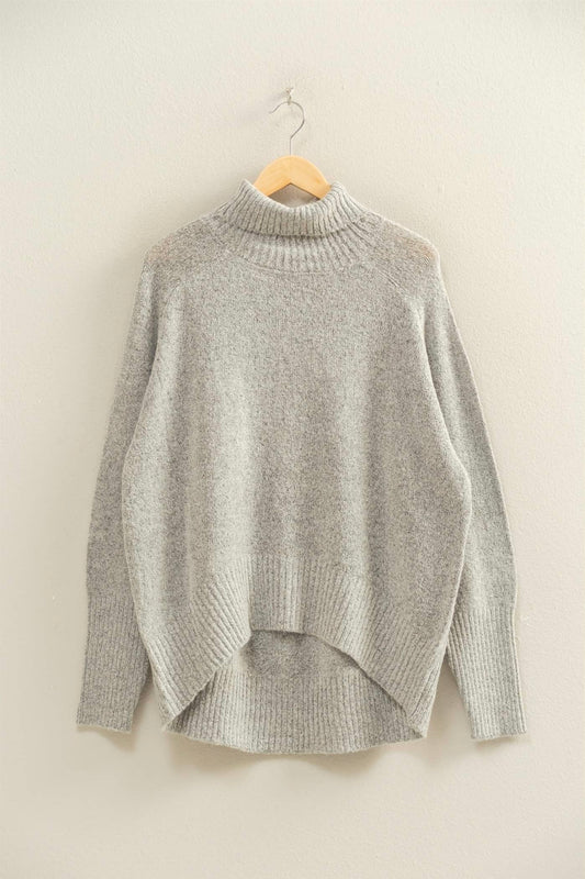 Heather grey mock-neck sweater hanging on a wooden hanger against a neutral wall. Ribbed neckline, cuffs, and hem with a relaxed silhouette and soft knit texture.