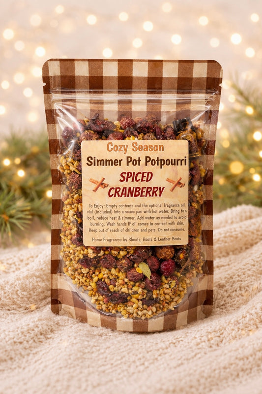 A stand-up resealable pouch of spiced cranberry simmer pot potpourri, shown on a soft beige fabric with blurred warm lights in the background. The pouch has a brown-and-beige gingham border and a cream label reading 'Cozy Season Simmer Pot Potpourri Spiced Cranberry.