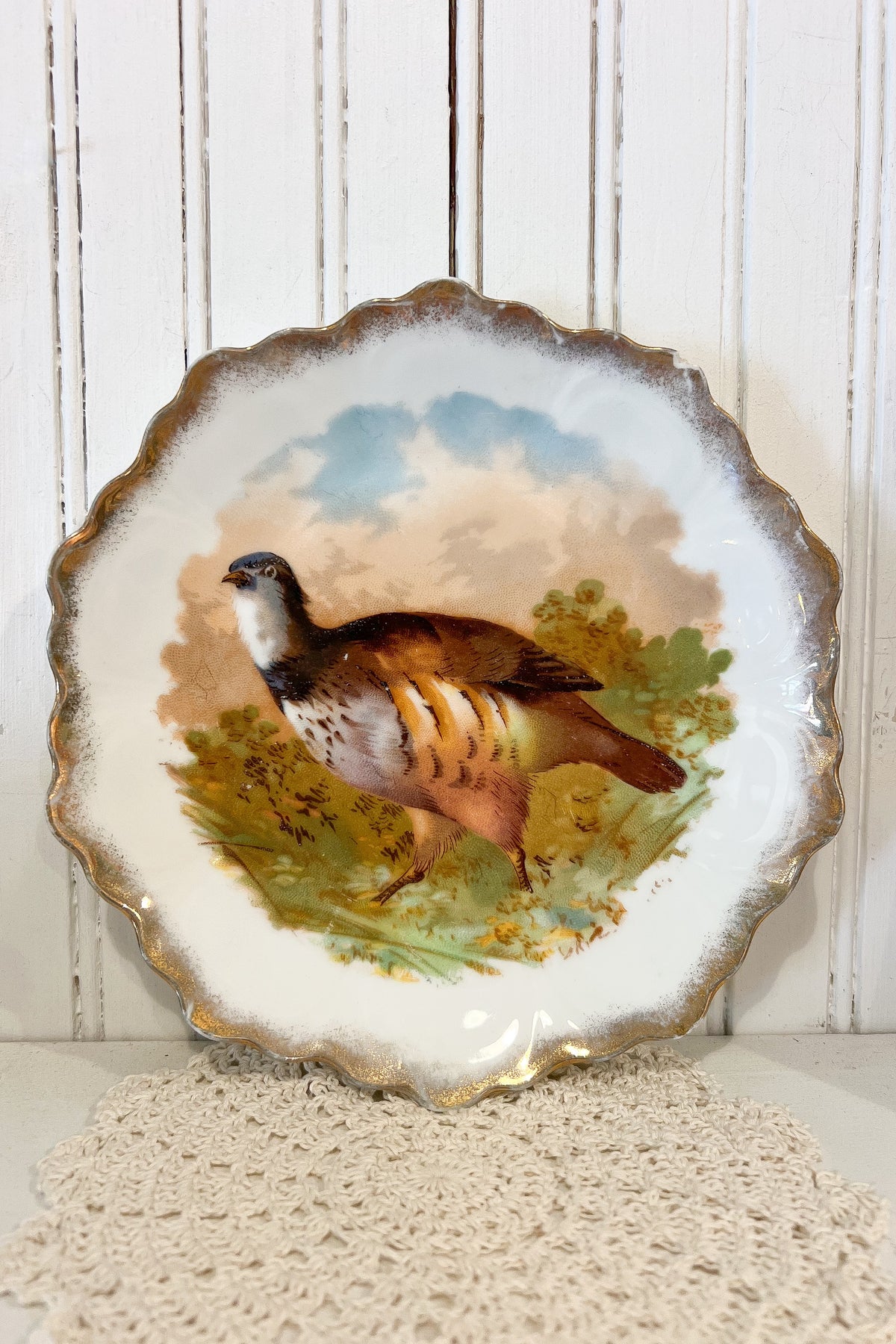 A scalloped ceramic plate featuring a hand-painted pheasant bird in a natural landscape with green foliage and a blue sky, displayed against a white wood-paneled background.