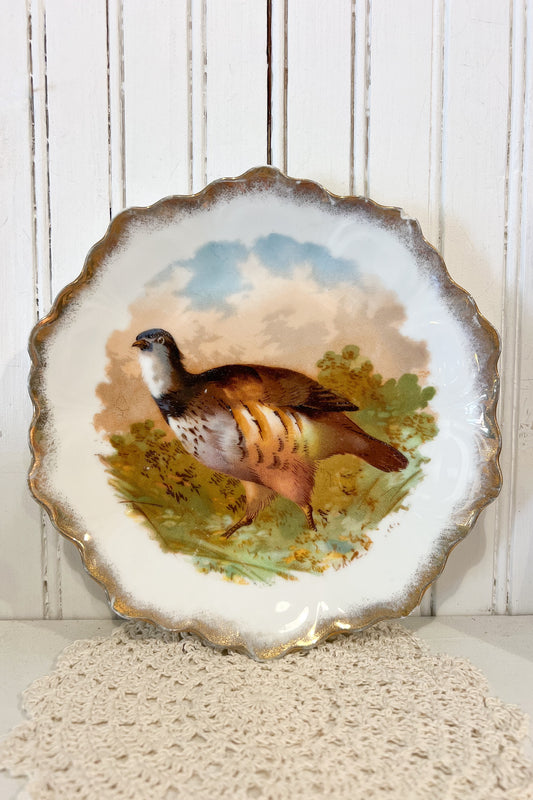 A scalloped ceramic plate featuring a hand-painted pheasant bird in a natural landscape with green foliage and a blue sky, displayed against a white wood-paneled background.