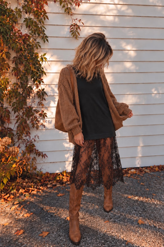 Model wearing a washed black short-sleeve tee dress with a sheer black floral lace midi layer, styled with a caramel cardigan, brown boots, and a tan shoulder bag.