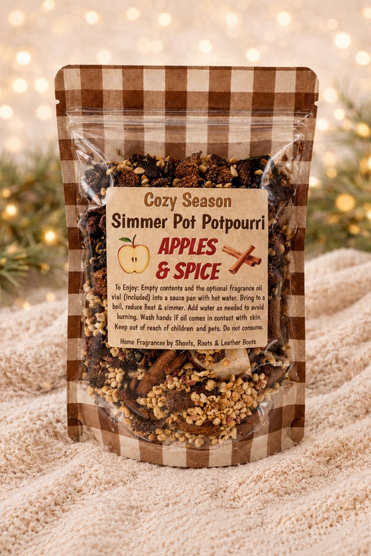 A stand-up resealable pouch of 'Cozy Season Simmer Pot Potpourri' by Home Fragrance by Sheets & Leather Boots, featuring a brown and beige gingham pattern and a central label with an apple and cinnamon graphic. The product is displayed on a soft beige knit blanket with a warm, blurred bokeh background.