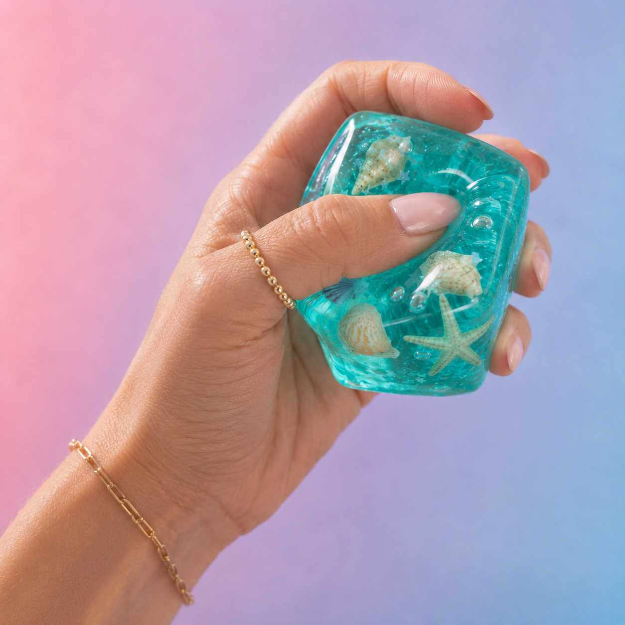 A hand holding a translucent turquoise squishy cube filled with seashells and a starfish, against a pink-to-blue gradient background.