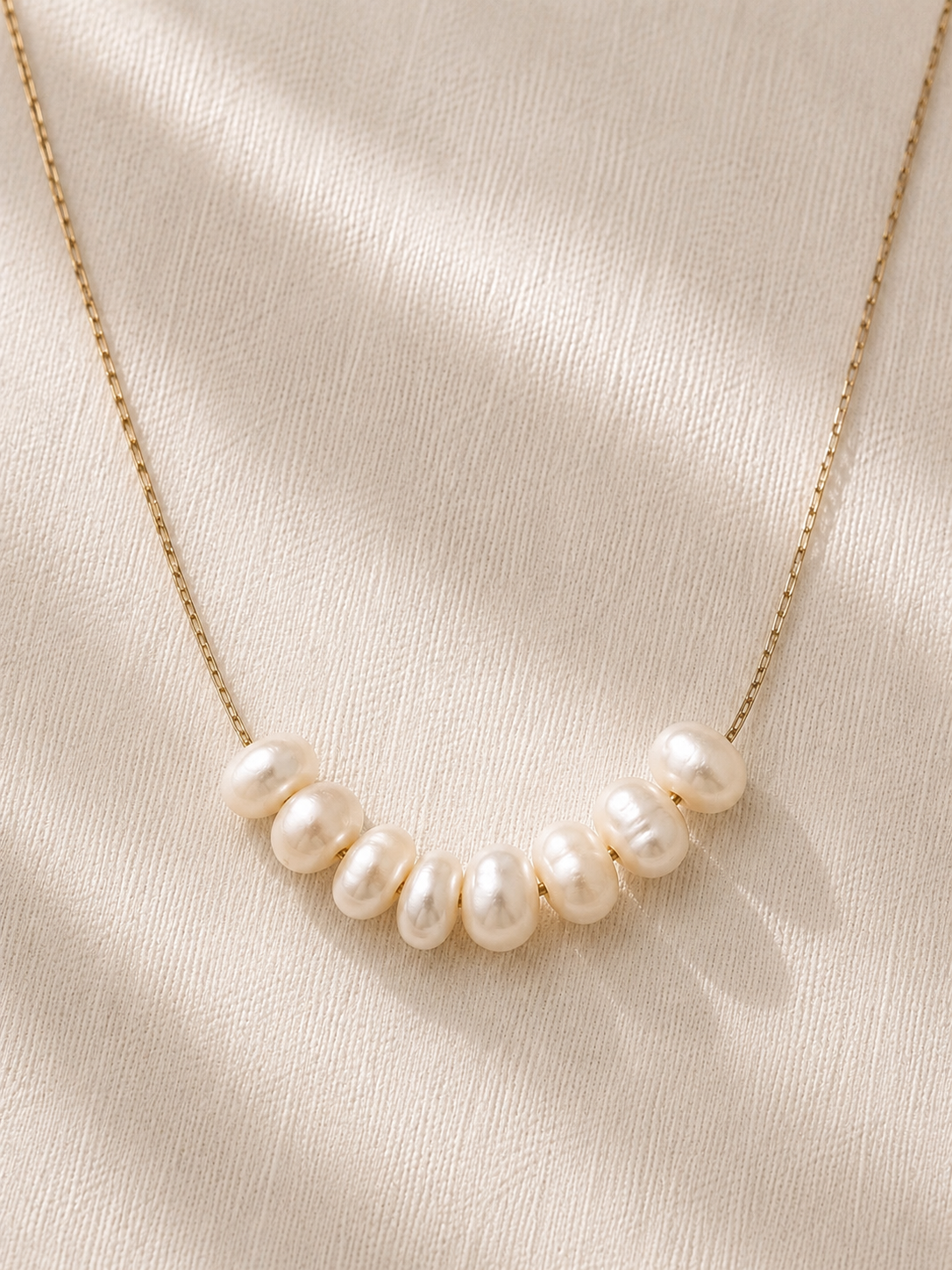 Gold necklace featuring a row of organically shaped freshwater pearls displayed on soft ivory linen.