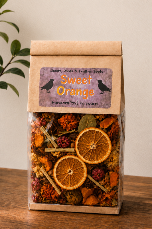 A clear plastic bag with a kraft paper top and a purple label reading 'Sweet Orange Handcrafted Potpourri,' filled with dried orange slices, flowers, and bark, standing on a wooden surface against a light wall.