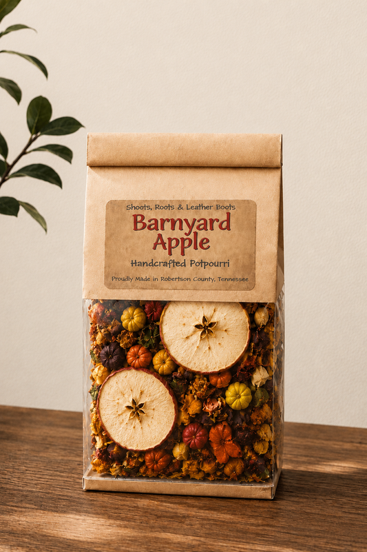 A kraft paper bag of Barnyard Apple handcrafted potpourri sits on a wooden table against a neutral beige wall. The clear lower section of the bag reveals a colorful mix of dried flowers, pinecones, and apple slices.