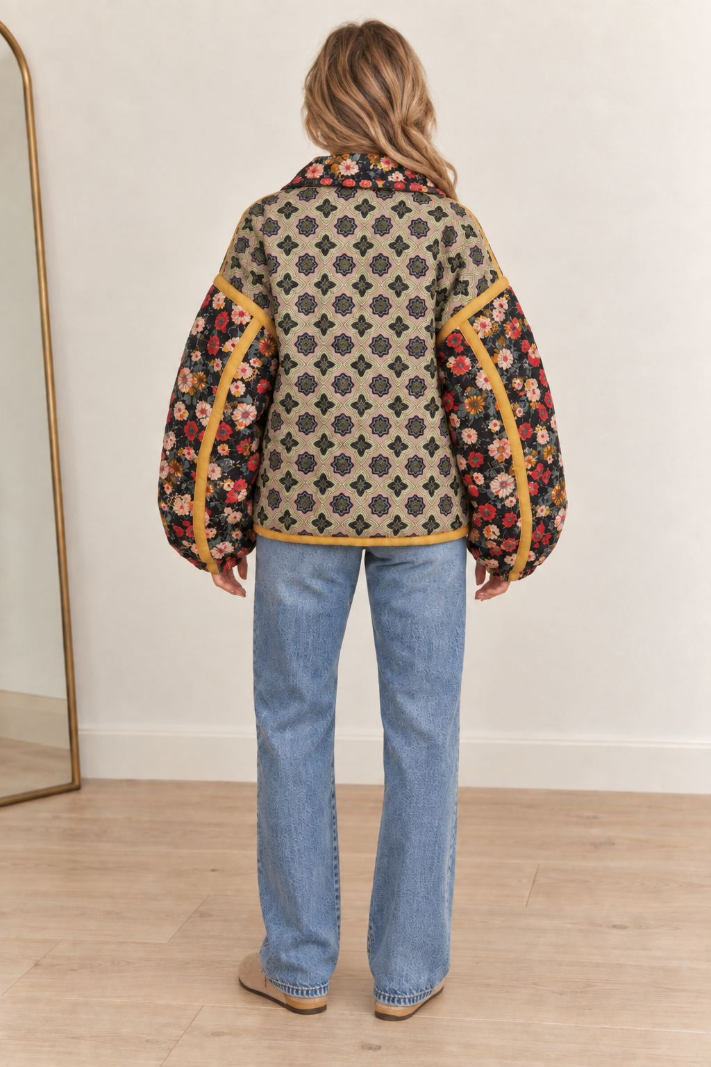 A woman viewed from behind wearing a quilted jacket with a beige and black geometric pattern and floral sleeves, paired with light blue jeans in a minimalist room with a wooden floor and a mirror.