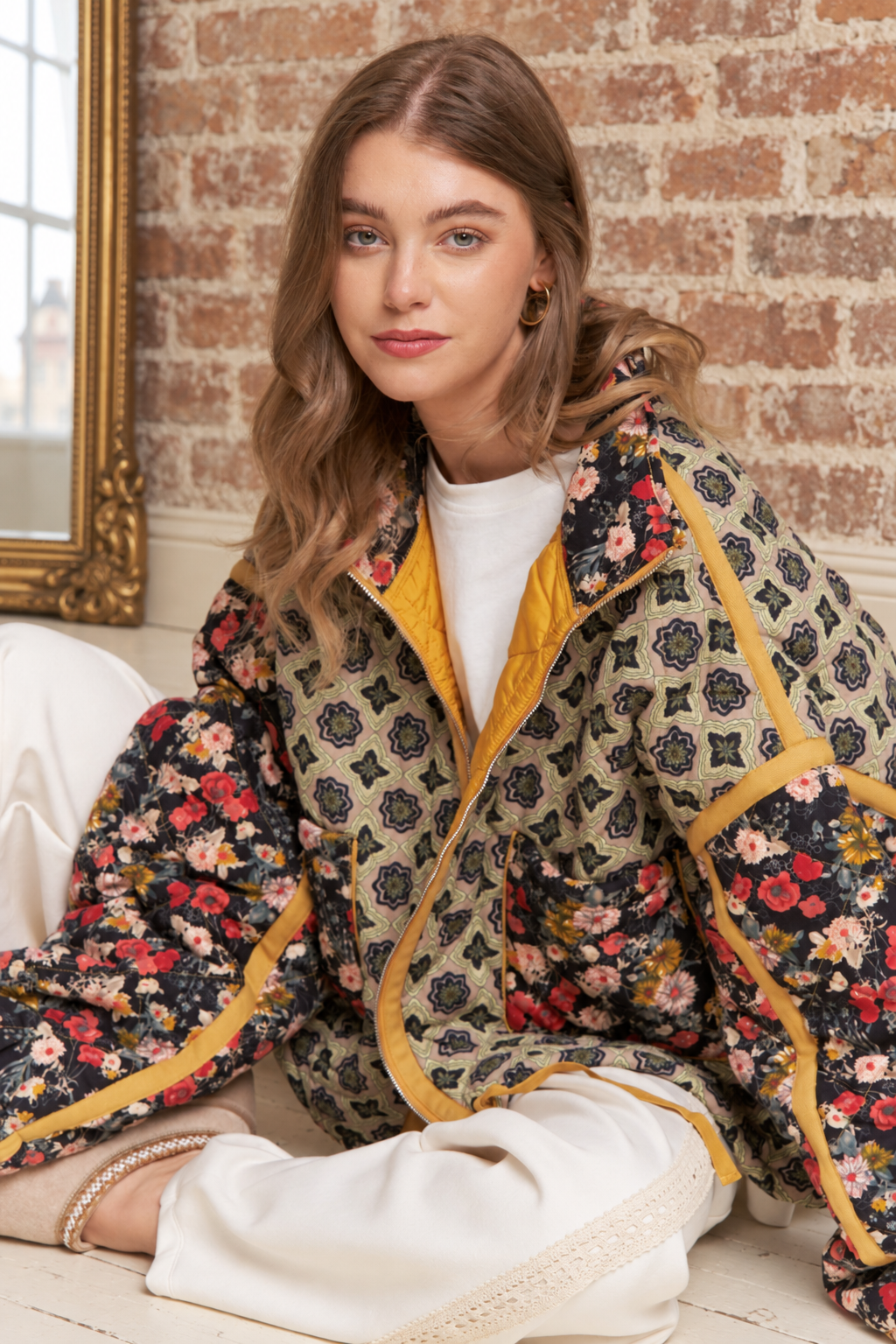A woman wearing a patterned quilted jacket with a floral print, yellow lining, and white pants, sitting in front of a brick wall.