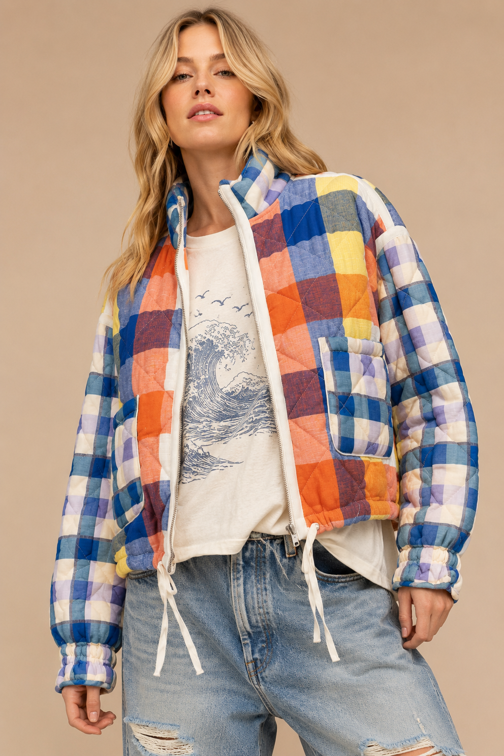 A woman models a colorful patchwork quilted jacket with blue, orange, yellow, and purple gingham panels, worn over a white graphic t-shirt and light-wash distressed jeans against a solid beige background.