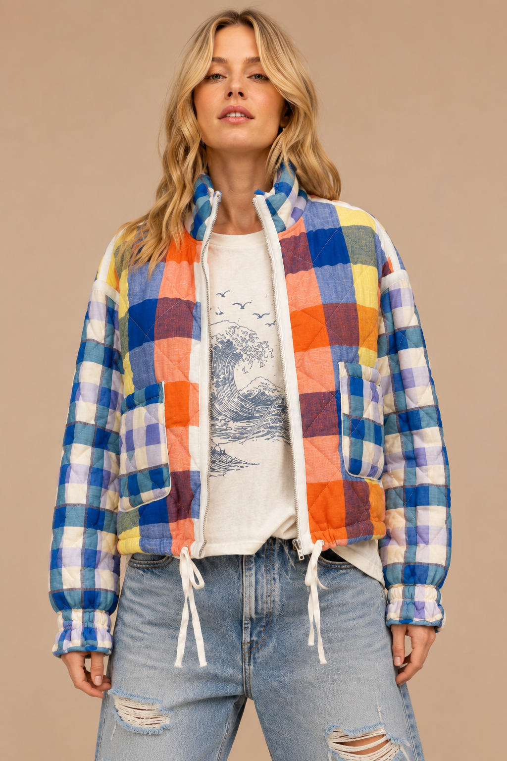 A woman modeling a colorful patchwork quilted jacket with blue, yellow, orange, and purple plaid panels, worn open over a white graphic t-shirt and light-wash distressed jeans against a solid beige background.