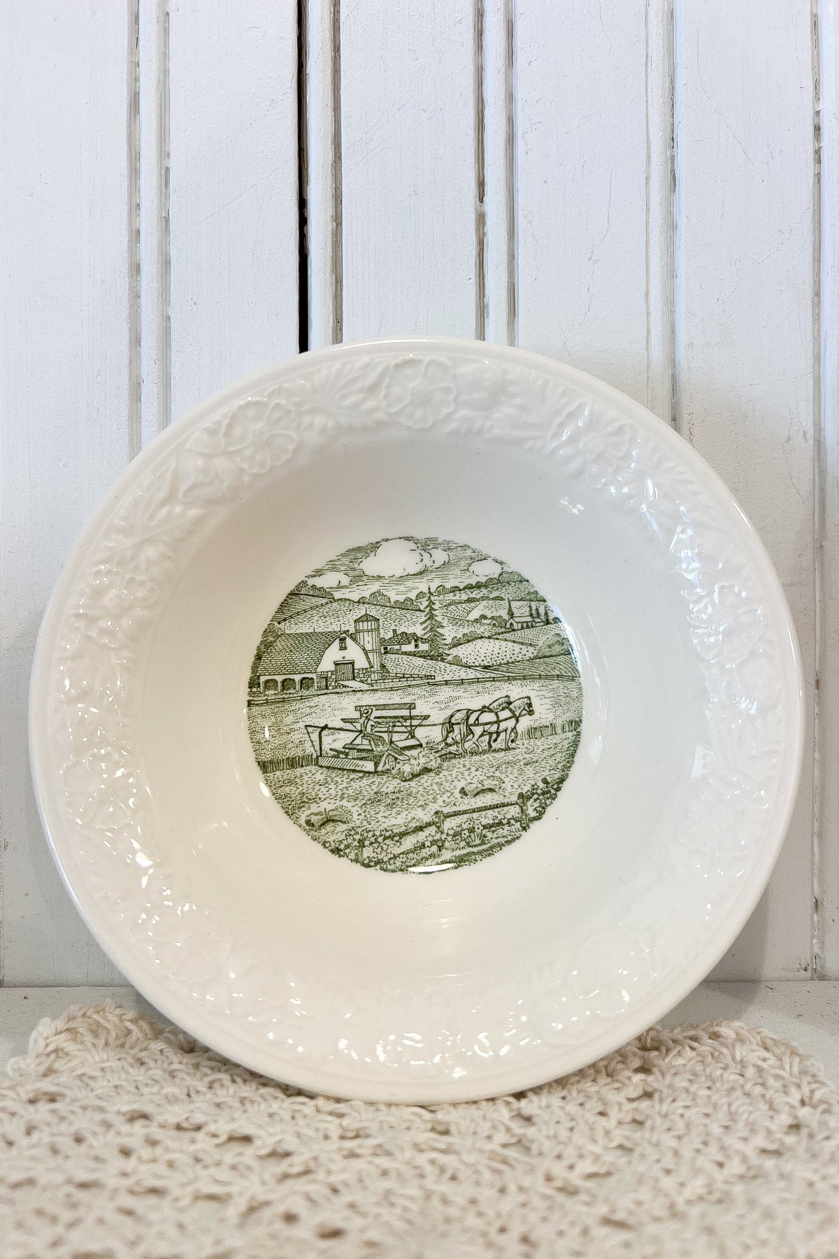 A white ceramic bowl with an embossed floral pattern on the rim and a green landscape illustration printed in the center, displayed on a textured cream doily against a white wooden paneled background.
