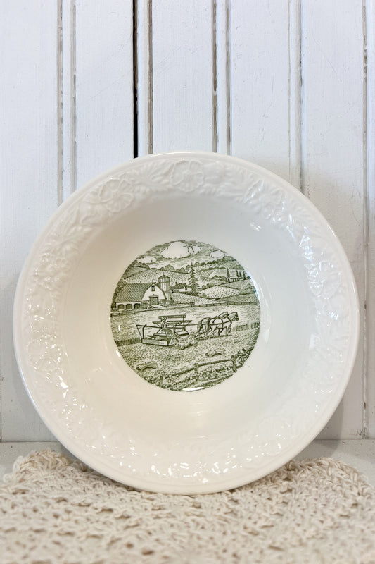 A white ceramic bowl with an embossed floral pattern on the rim and a green landscape illustration printed in the center, displayed on a textured cream doily against a white wooden paneled background.