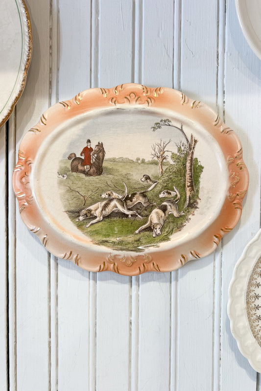 An oval vintage ceramic serving platter with a peach-colored scalloped rim and a detailed hunting scene in the center, displayed on a white painted wooden plank surface.