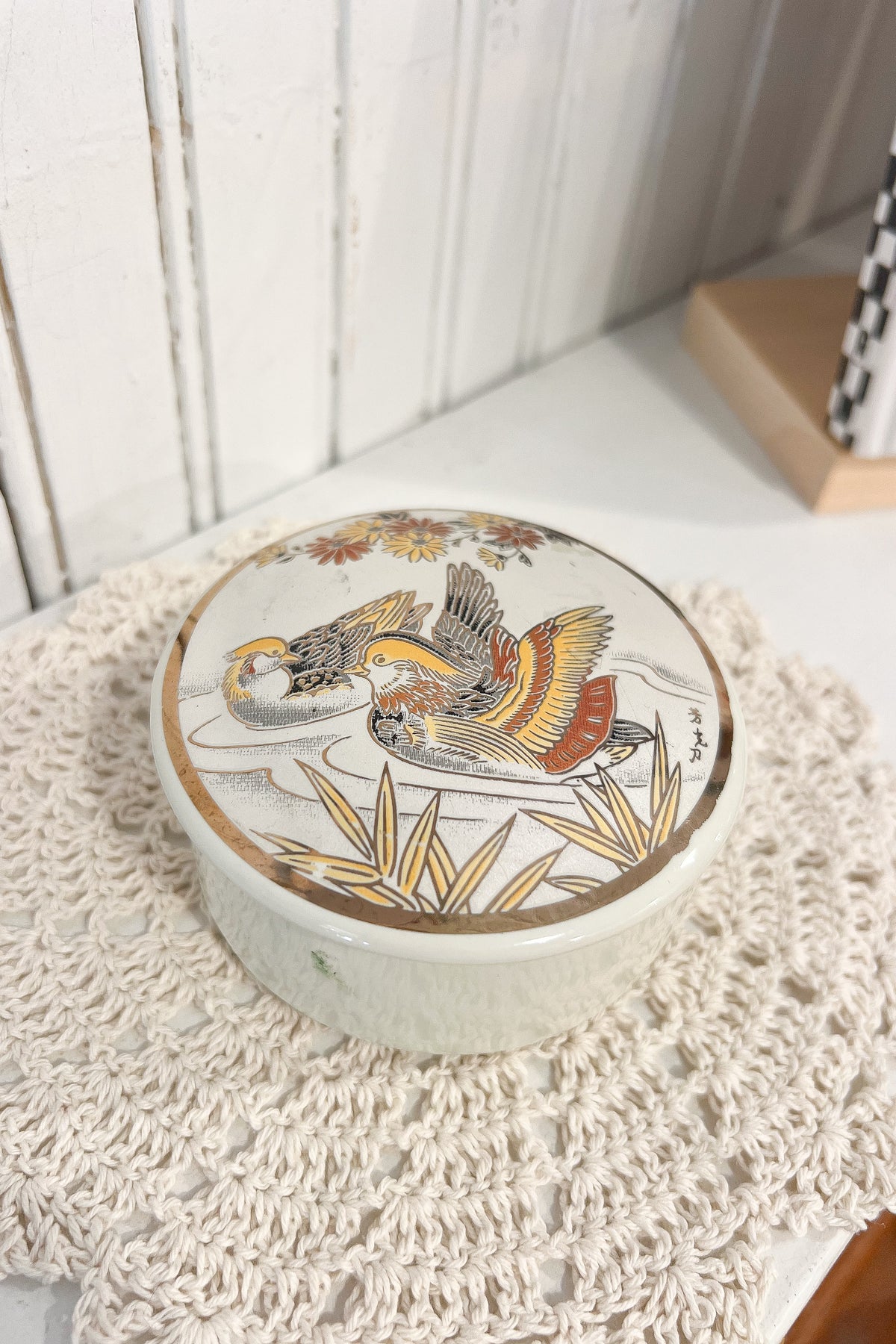 A round ceramic lidded box with a hand-painted Japanese-style crane design on the lid, resting on a cream-colored crocheted doily against a white paneled background.