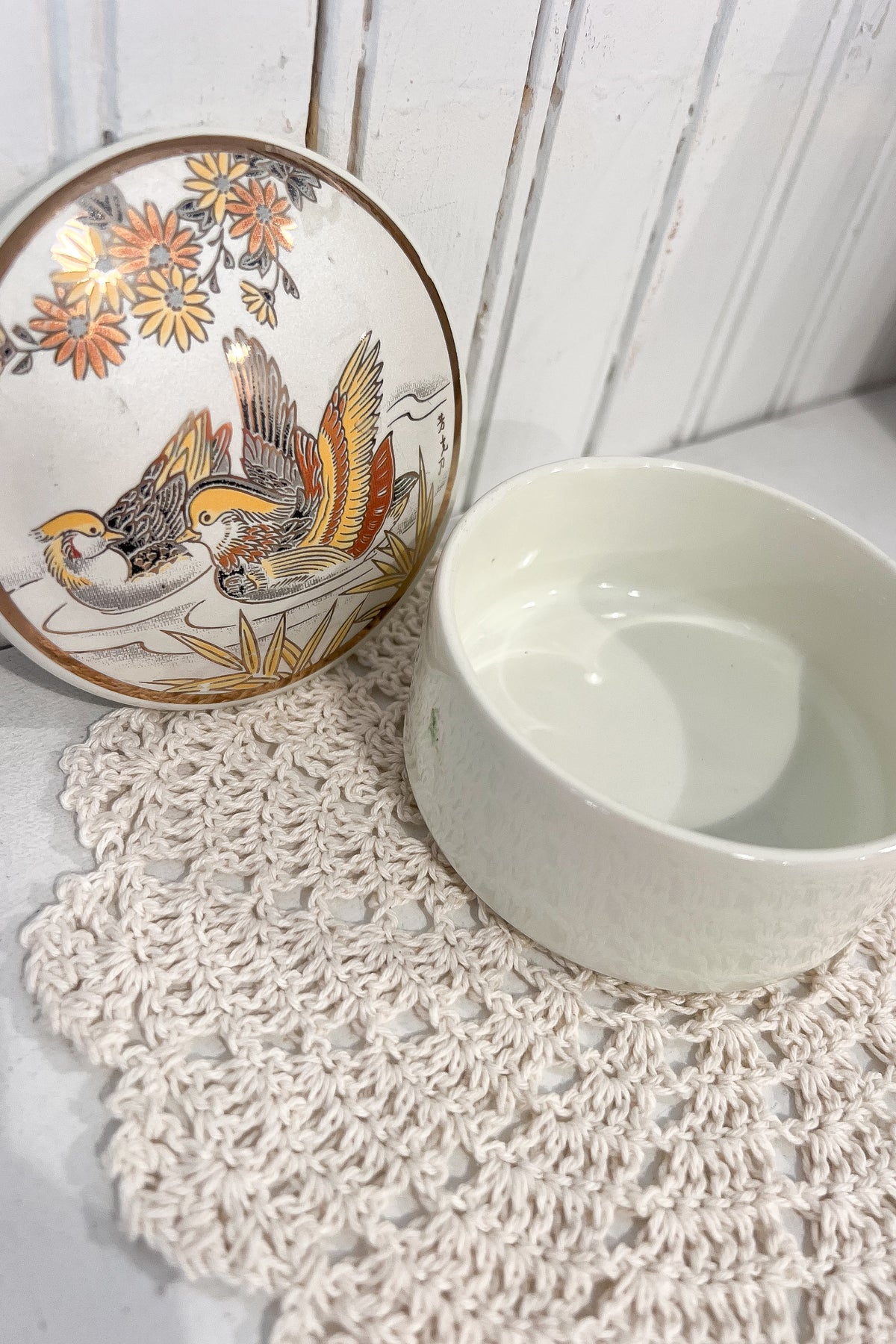 A white ceramic bowl with a textured exterior and a matching floral-patterned plate with a bird illustration, styled on a cream crochet doily against a white wood-paneled background.