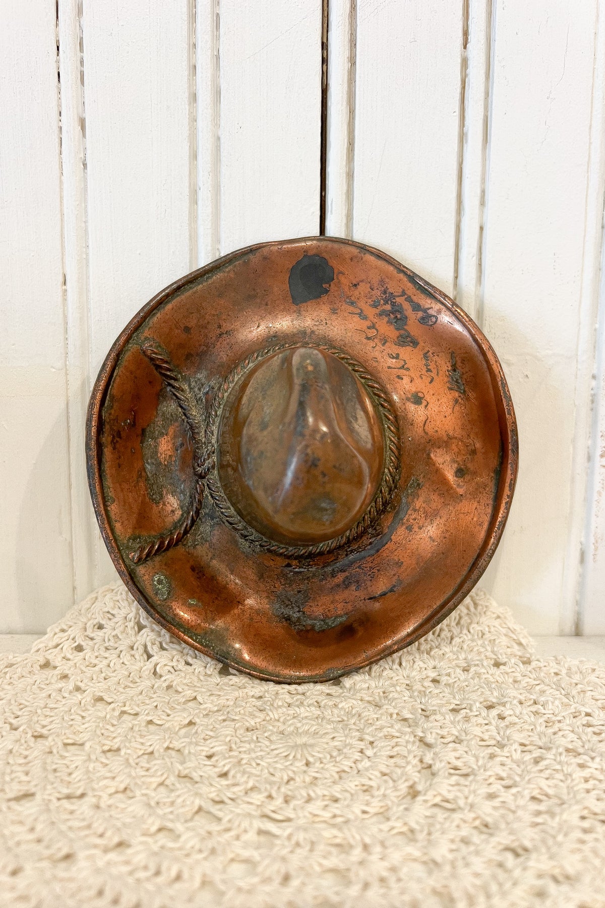A rustic, hand-hammered copper-colored metal hat-shaped plate or tray with a distressed, weathered finish, displayed on a cream-colored textured fabric against a white wooden paneled background.