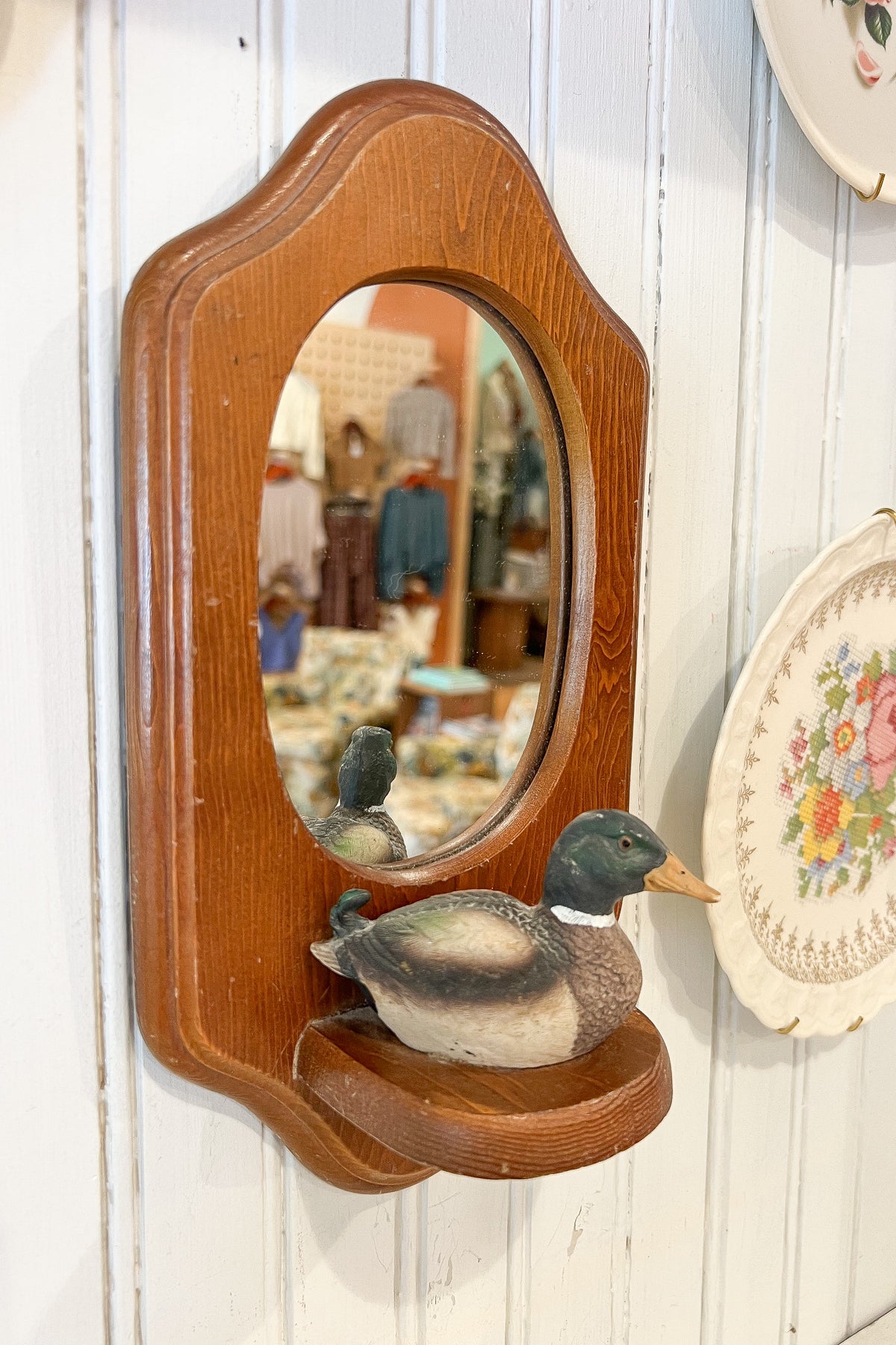A vintage-style oval wooden wall mirror with a carved duck-shaped base, mounted on a white paneled wall. The mirror reflects a shop interior, and a decorative floral plate is partially visible on the right.