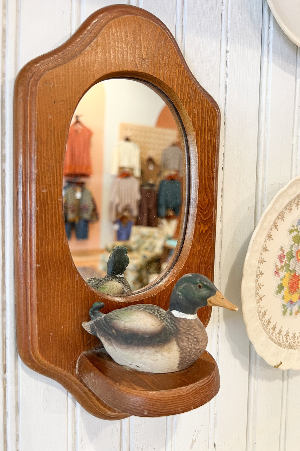A vintage-style wooden wall mirror with an oval reflective center and a small wooden shelf below, styled with a duck figurine on the shelf. The mirror is mounted on a white paneled wall, with a decorative plate partially visible on the right.