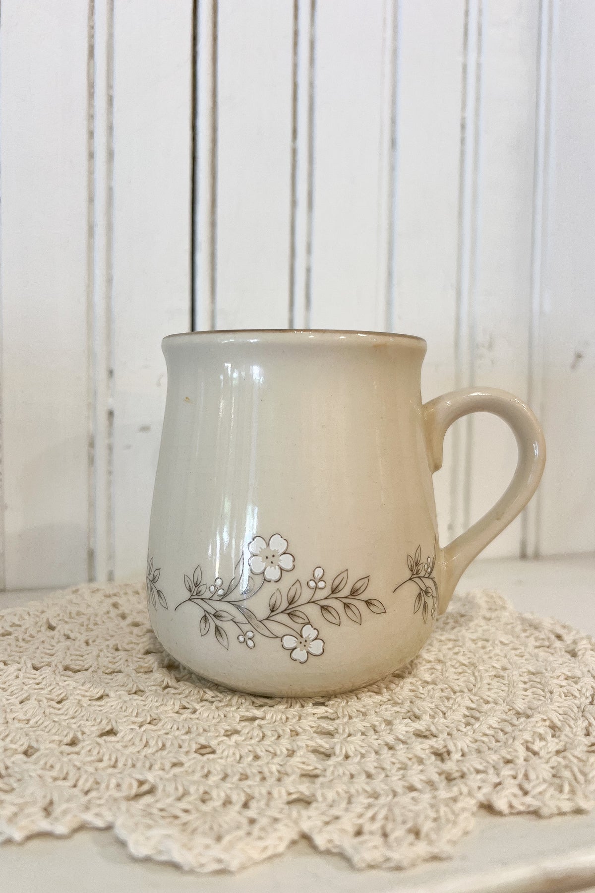 A cream-colored ceramic mug with a delicate floral vine pattern, resting on a white crocheted doily against a white wooden paneled background.