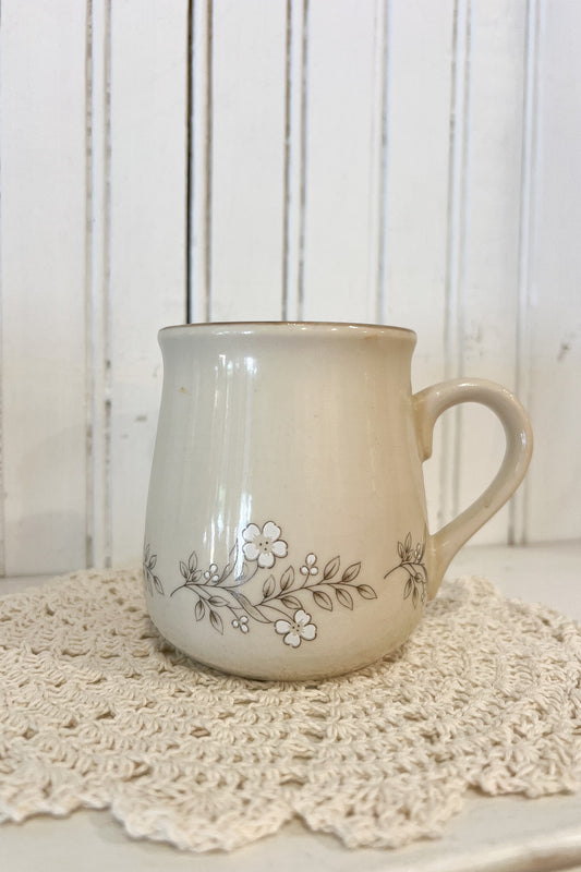 A cream-colored ceramic mug with a delicate floral vine pattern, resting on a white crocheted doily against a white wooden paneled background.