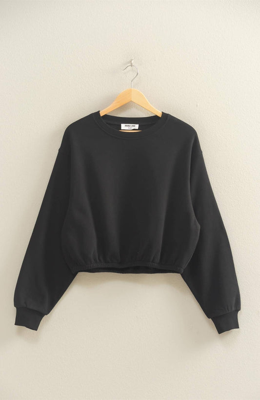 A black cropped cinch-hem sweatshirt hanging on a wooden hanger against a light wall. Minimal, cozy, and modern — perfect for everyday wear or loungewear styling.