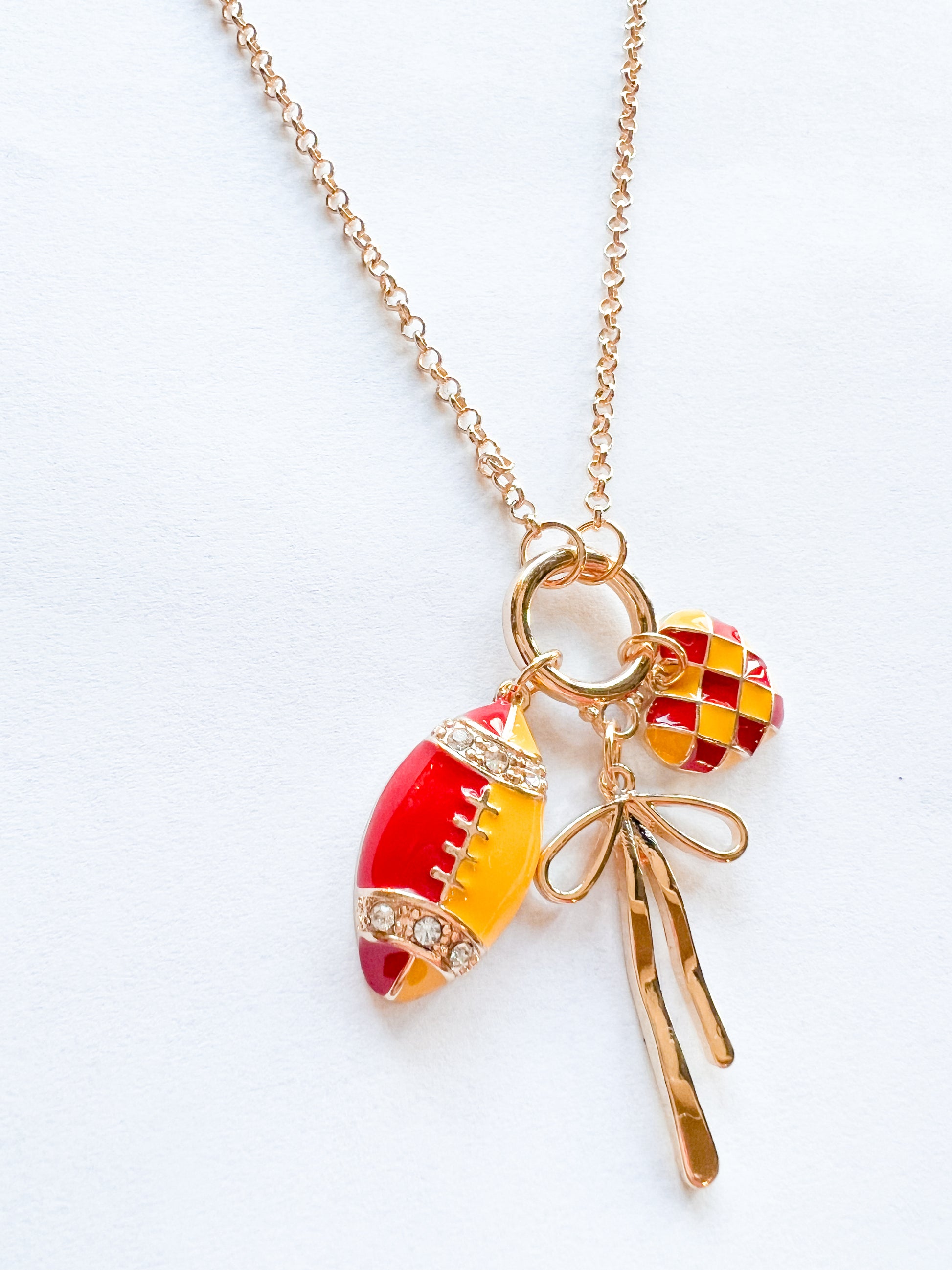 Gold chain necklace with red and yellow football charm, checker heart charm, and delicate gold bow—KC Chiefs inspired game day jewelry on white background.