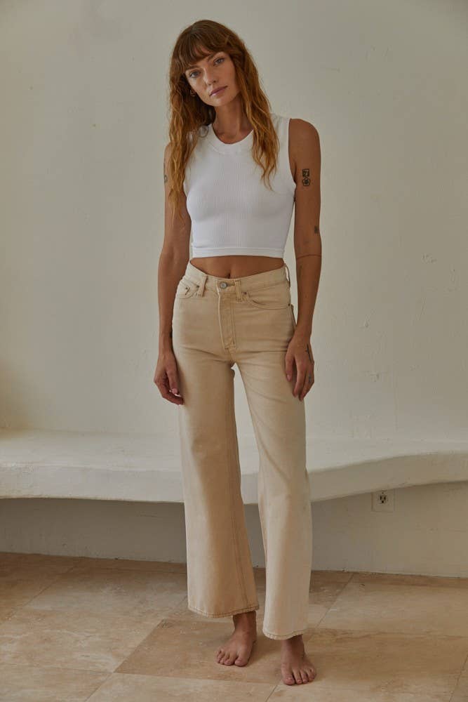 Woman wearing a white tank top and beige pants standing against a plain wall.