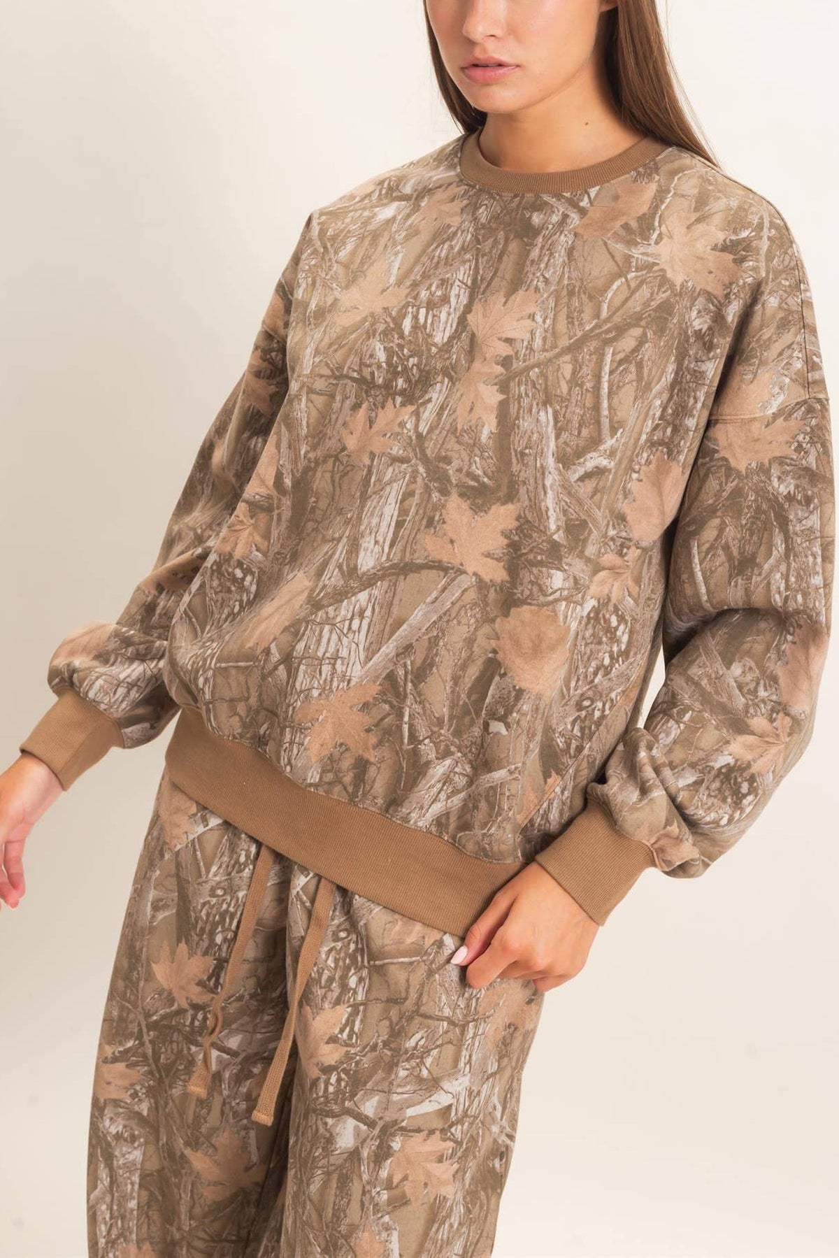 A woman modeling a matching camouflage sweatshirt and sweatpants set in a light studio setting. The camouflage pattern features shades of brown, beige, and tan, with solid brown ribbed cuffs and a matching brown waist tie.
