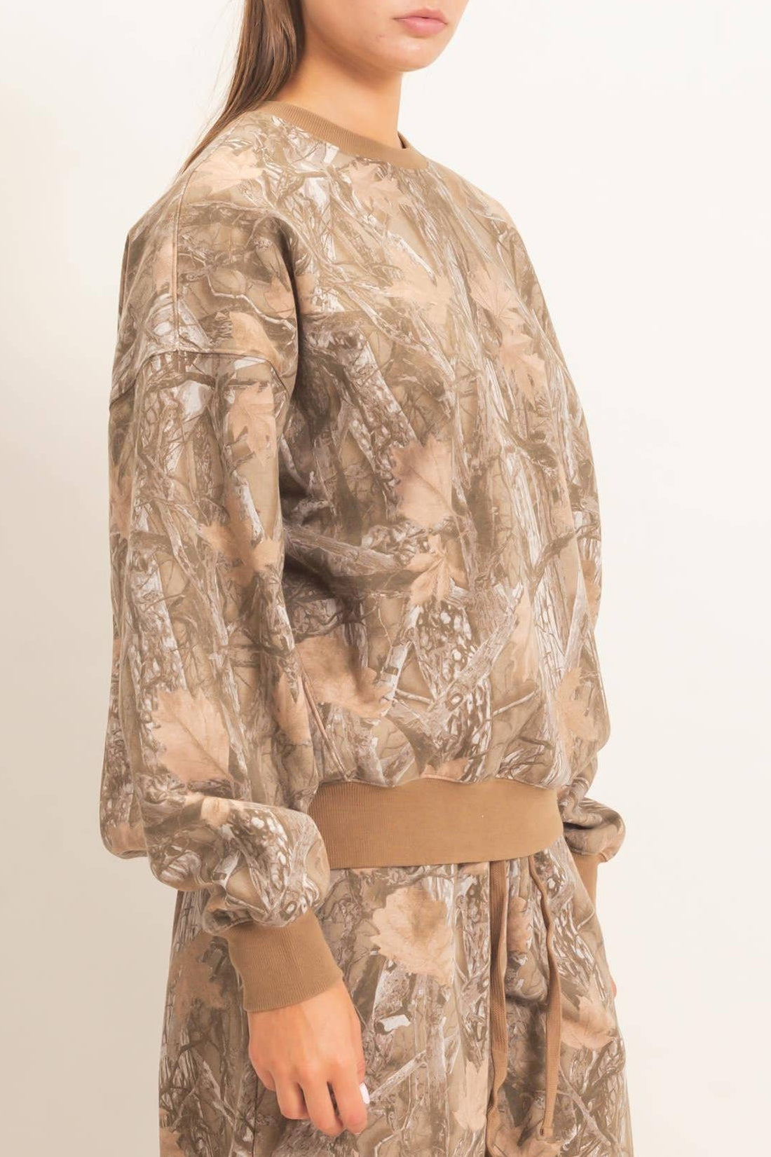 A woman modeling a matching camouflage sweatshirt and sweatpants set in a light brown and beige woodland camo pattern, photographed against a plain off-white studio background.