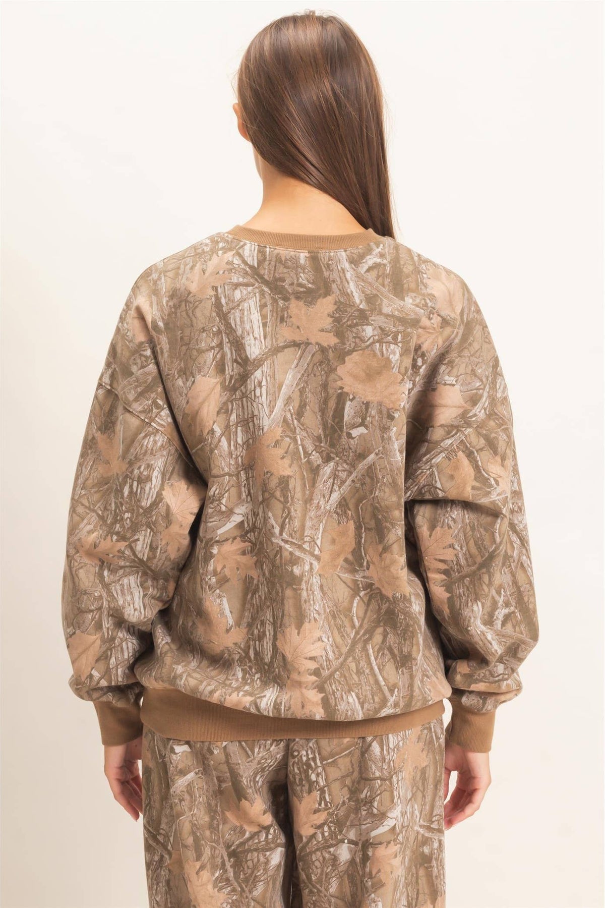 A woman shown from the back wearing a brown and beige camouflage sweatshirt and matching pants against a plain off-white studio background.