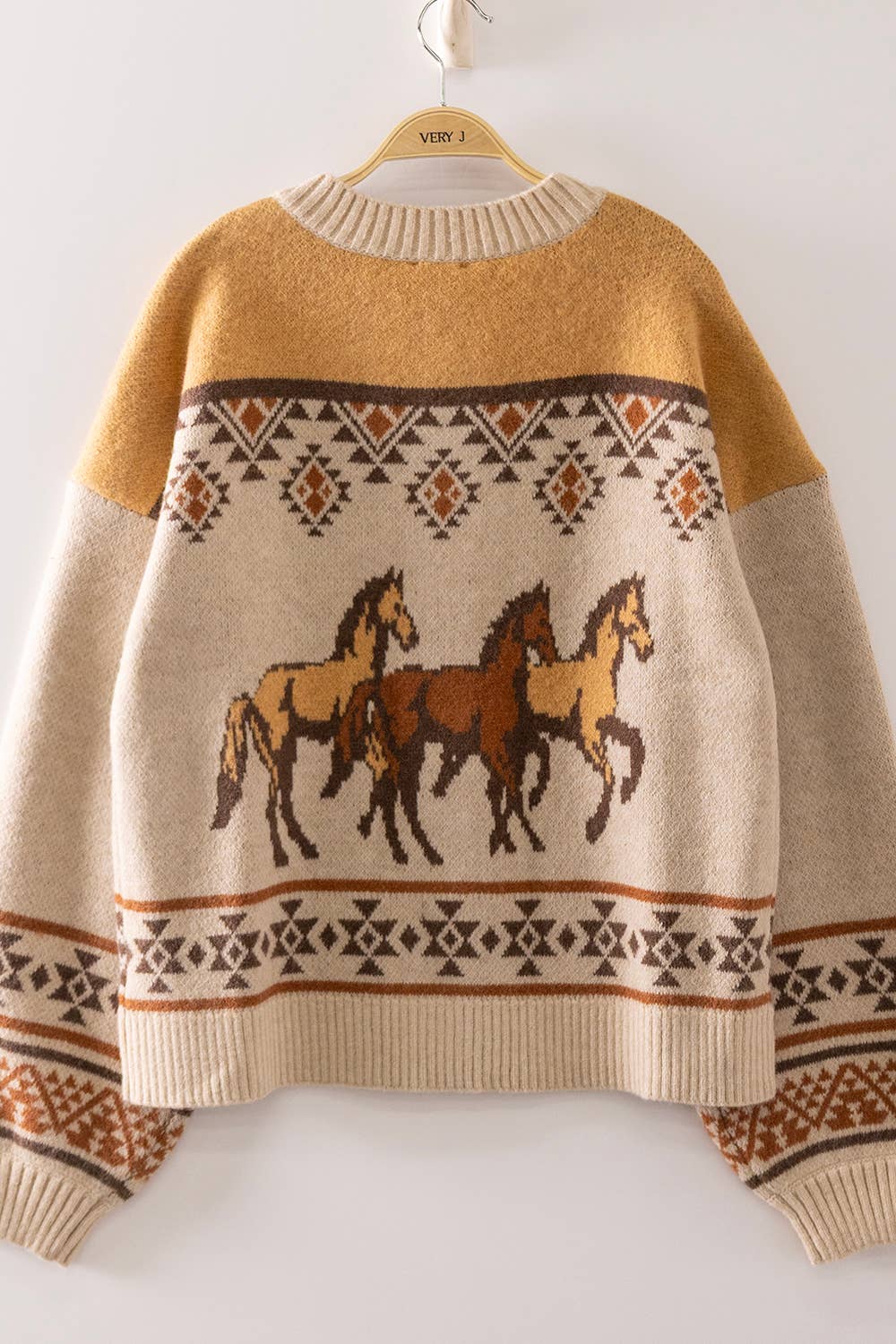A knit crewneck sweater hanging on a wooden hanger against a plain light background. The sweater has a mustard yellow upper yoke, a cream lower body, and a central pattern featuring three horses in brown and tan, bordered by geometric tribal-style patterns.