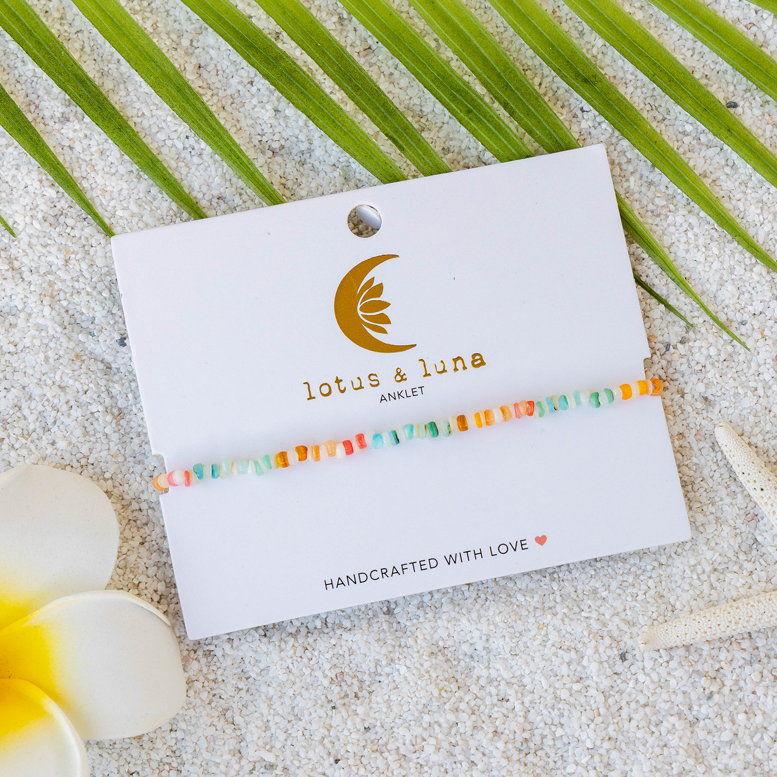 Colorful beaded anklet on a 'lotus & luna' card with sand and palm leaves in the background